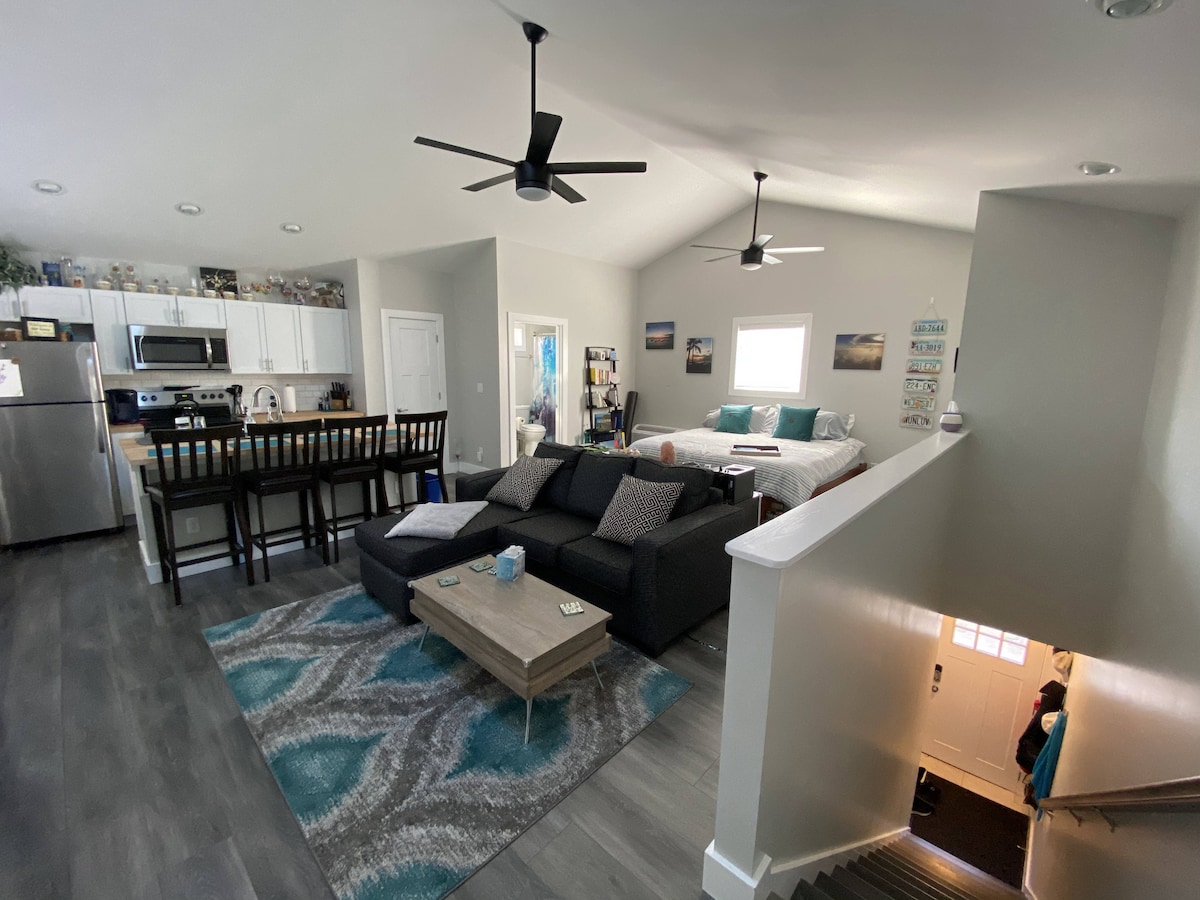 An open-concept living area features a spacious sectional sofa adorned with patterned throw pillows. A wooden coffee table sits on a decorative rug. The kitchen area includes a bar with seating, and a staircase leads to the entrance, showcased by natural light filtering through nearby windows.