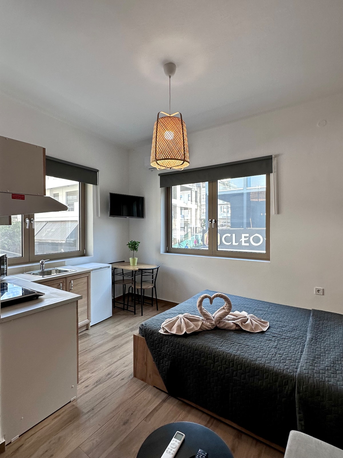 A compact room features a spacious bed with a textured dark blanket, complemented by a decorative towel arrangement. A small dining table with two black chairs sits near the window, which provides natural light and a view of the street. A flat-screen TV is mounted on the wall.