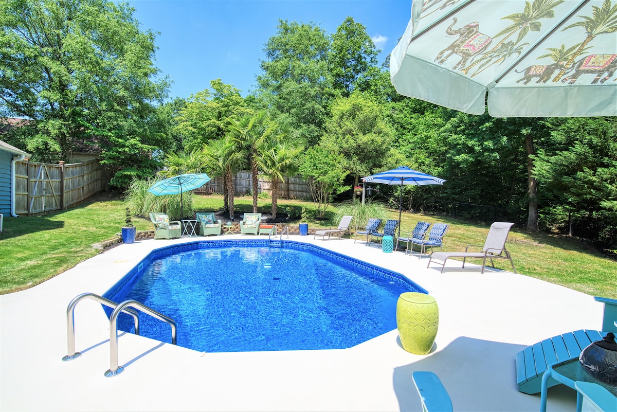 A tranquil outdoor space features a shimmering in-ground saltwater pool surrounded by lush greenery. Lounge chairs, umbrellas, and a bright green accent table provide comfort and shade. The fenced yard creates a private oasis, ideal for relaxation or gatherings.
