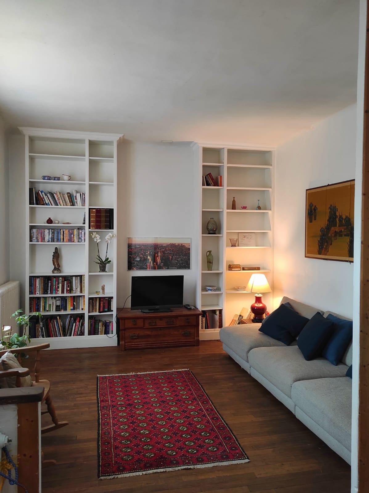 A cozy living room is presented, featuring a soft sofa adorned with decorative pillows. A wooden cabinet holds books, while a small television sits on a stand. Wall shelves display various items, and warm light from a lamp adds to the home's inviting atmosphere.