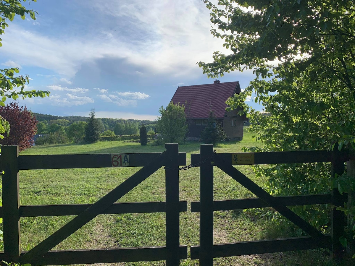 A wooden cabin is visible beyond a rustic gate, surrounded by lush greenery. The site is framed by trees, with a clear sky above hinting at a peaceful atmosphere. The grassy field extends towards the horizon, creating an inviting view of the natural surroundings.