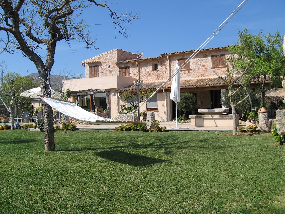 A charming stone house is set in a tranquil garden, featuring a well-maintained lawn and a hammock tied between trees. The exterior showcases a blend of traditional architecture and natural elements under a clear blue sky, inviting relaxation and outdoor enjoyment.
