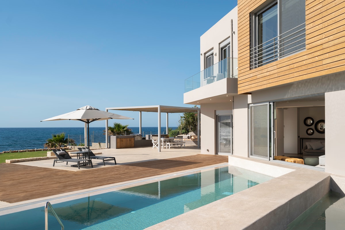 A modern villa overlooks the ocean, featuring a sparkling private pool and wooden deck. Comfortable lounge chairs are positioned beneath a large umbrella, while an outdoor dining area is shaded nearby. The expansive windows invite natural light into the stylish interior.