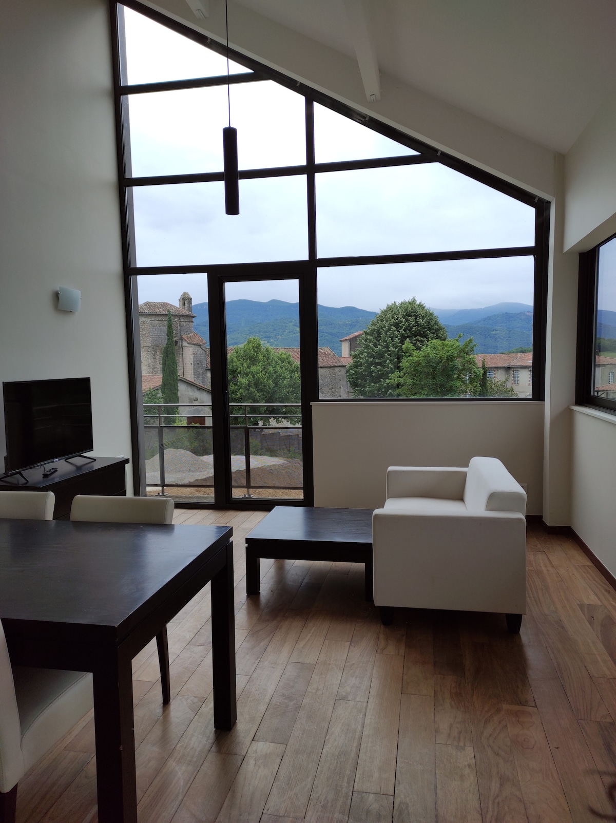 A bright living area features large windows, offering views of the surrounding village and mountains. A wooden table and chairs provide dining space, while a light-colored sofa and coffee table create a comfortable seating arrangement. Natural light enhances the modern interior design.