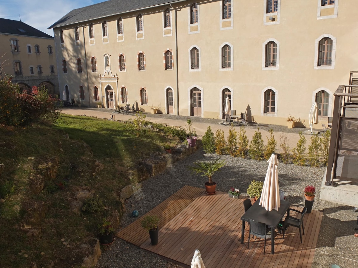 An outdoor terrace features a wooden deck with a dining table and chairs, surrounded by potted plants and umbrellas. The view includes the historic building facade, showcasing multiple windows and a well-maintained garden area with gravel pathways.