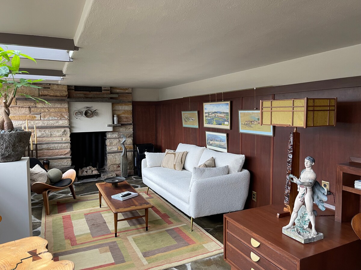 A main living area is featured with a light-colored sofa and a wooden coffee table. Stone walls surround a fireplace, complemented by wooden panels and framed art. A colorful rug lies beneath, while a stylish lamp and greenery add a touch of warmth.