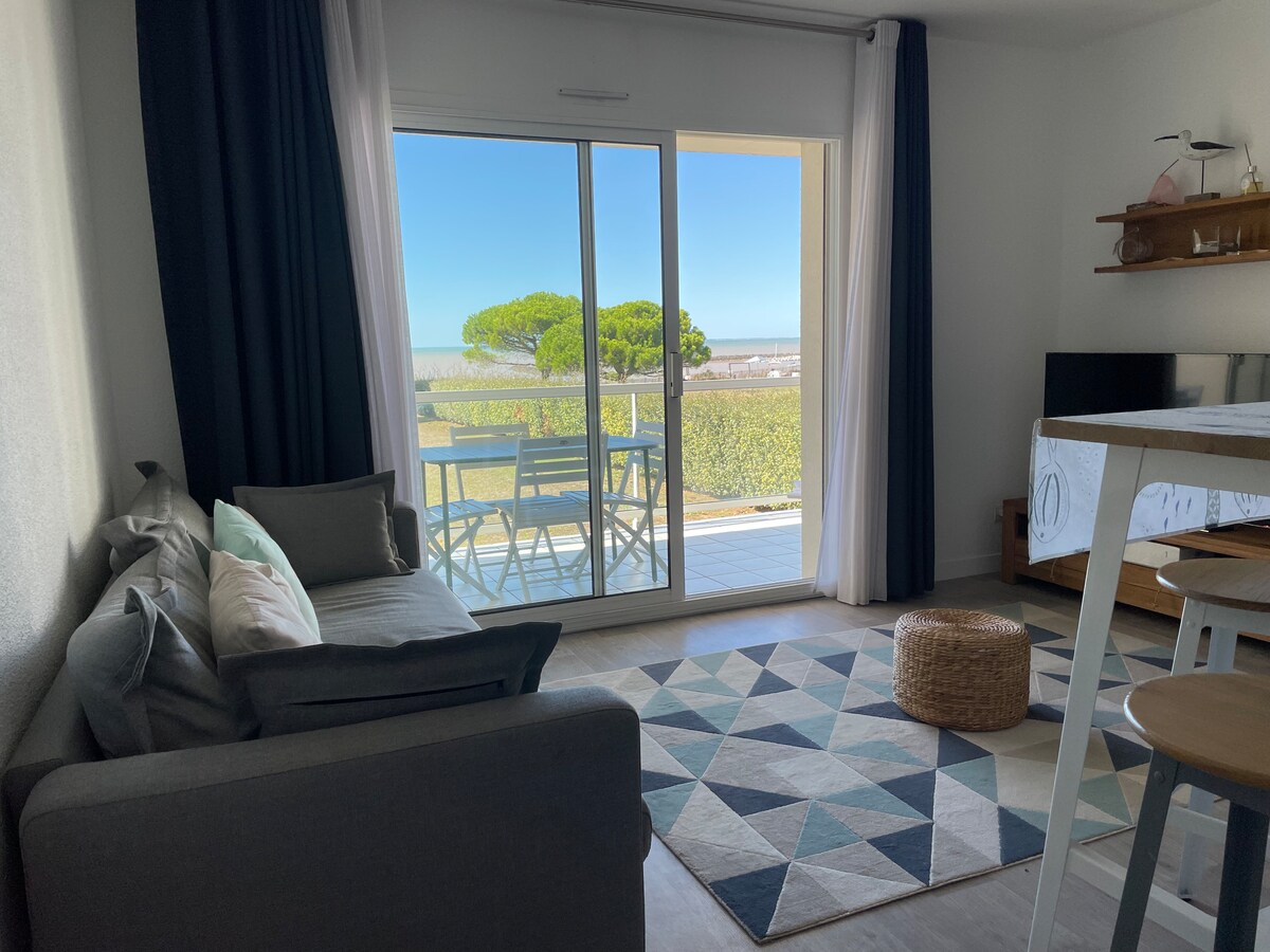 A bright living area is featured, with a light grey sofa facing sliding glass doors that open to a balcony. A small dining table with four chairs and a patterned rug are visible. Soft blue curtains frame the large windows, offering a glimpse of outdoor greenery.
