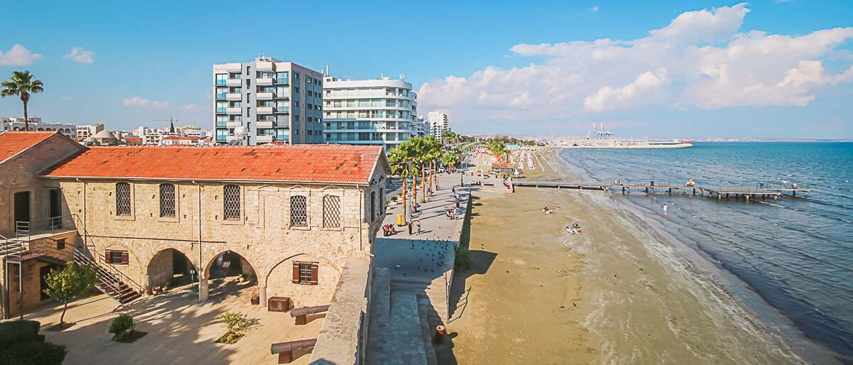 The image captures a scenic view of the coastline along the beach. A sandy shore stretches out next to low tide, while people are seen enjoying the water. A promenade lined with palm trees and modern buildings runs parallel to the beach, creating a vibrant urban atmosphere.