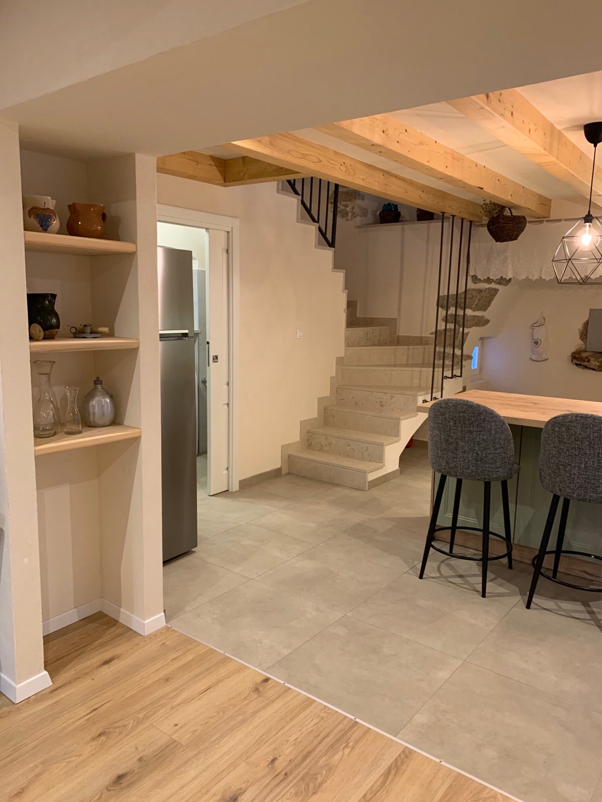 The image showcases an open kitchen and dining area with modern finishes. A staircase leads to the upper floor, while an island with seating is positioned near the kitchen. Shelving units are visible, displaying decorative items and kitchenware.