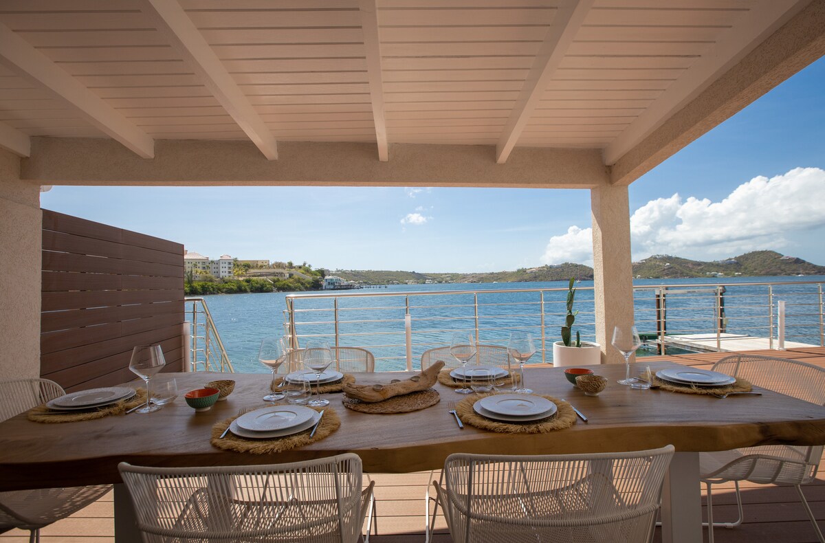 A spacious outdoor dining area features a long wooden table set with plates and glasses. Surrounding chairs offer comfortable seating. The view of the lagoon is unobstructed, with water reflecting the blue sky and distant land features lining the horizon.