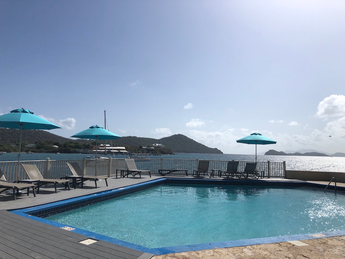 A refreshing pool area is presented with a clear blue water pool surrounded by lounge chairs. Large teal umbrellas provide shade. The picturesque backdrop features rolling hills and calm ocean waters under a bright sky.