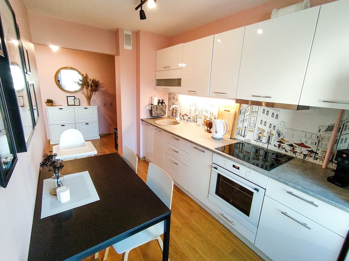 A modern kitchen is presented, featuring sleek white cabinetry and a light pink accent wall. A dining table with four chairs is positioned near the kitchen area. The countertop is equipped with cooking appliances, and a decorative mural of a café scene enhances the ambiance.