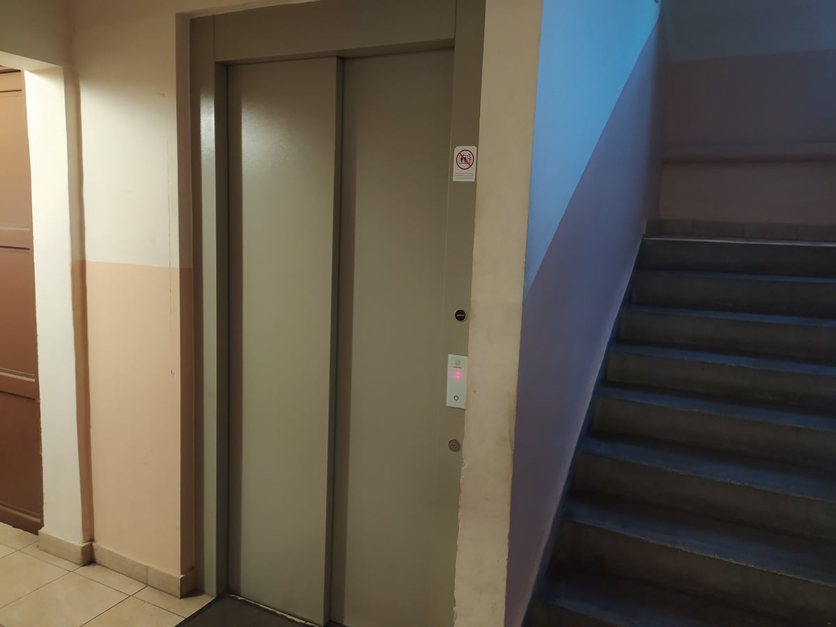 An elevator is positioned next to a staircase within a well-lit corridor. The elevator door is closed, and a call button panel can be seen, featuring a lit button indicating its operational status. Soft colors adorn the walls, contributing to a functional atmosphere.