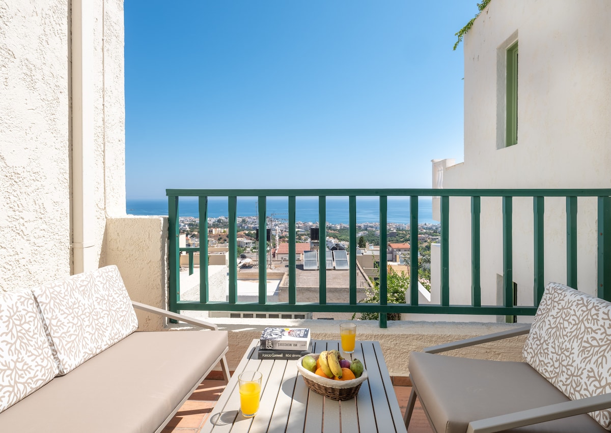 A balcony seating area features two comfortable lounge chairs and a coffee table, adorned with a fruit bowl. The space offers sweeping views of the sea and surrounding village, illuminated by bright sunlight, providing a serene environment for relaxation.