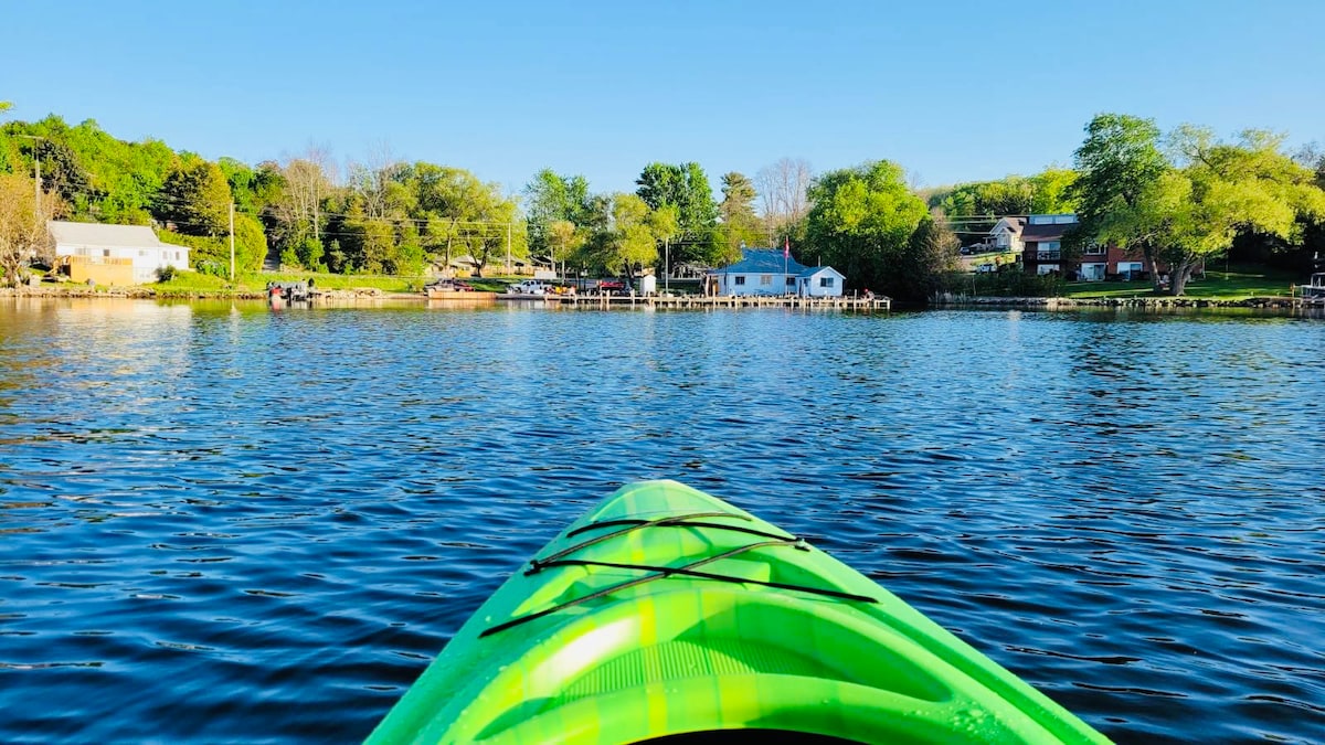 A vibrant green kayak is positioned at the forefront, facing a peaceful lake scene. The calm water reflects surrounding trees and cottages, while a few individuals can be seen enjoying leisure activities along the shore.