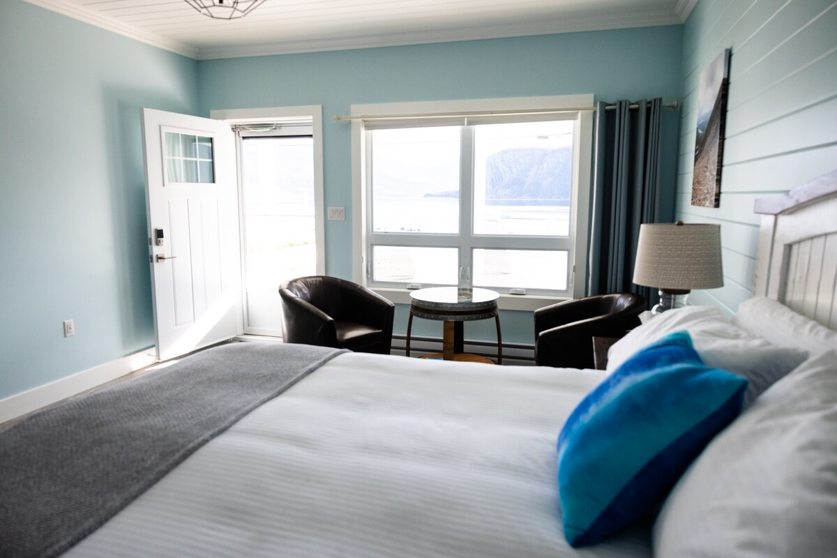A light-filled room features a spacious bed adorned with soft blankets. Two comfortable chairs are positioned near a round table by the window, providing views of the sea and mountains. A door leads to a private entrance, enhancing the room's inviting atmosphere.