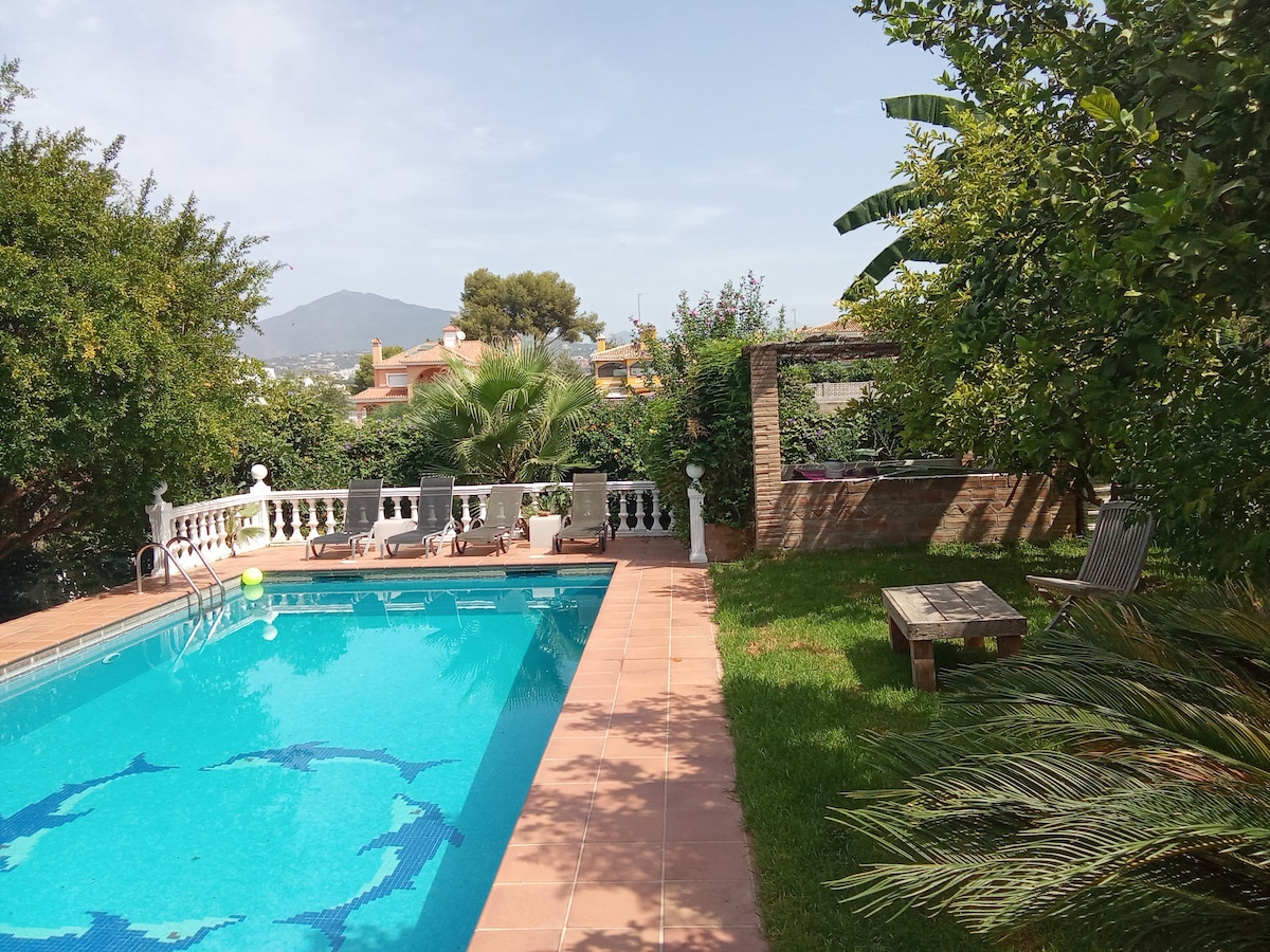 A serene outdoor space is highlighted, featuring a swimming pool surrounded by well-maintained gardens. Sun loungers are positioned on the poolside, and lush plants provide privacy. The backdrop includes a patio area with a shaded seating arrangement, enhancing the tranquil ambiance.