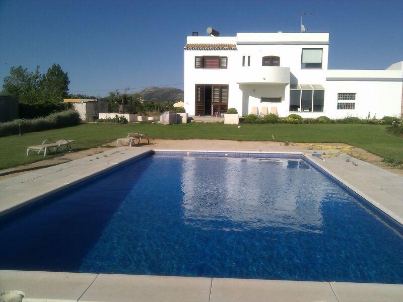 A spacious villa is shown, featuring a private swimming pool surrounded by neatly trimmed grass. Sun loungers are positioned along the poolside. The building offers a combination of large windows and a balcony, reflecting the bright blue sky and distant mountains.