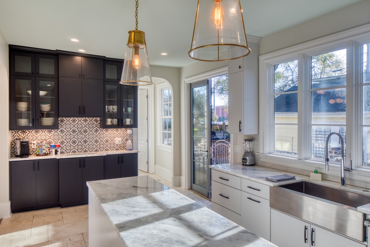 A spacious kitchen features a large island with Carrera marble countertop and contrasting cabinetry. Light streams through expansive windows, highlighting decorative backsplash tiles. Modern appliances, including a sink and blender, are present, while stylish pendant lights provide illumination over the island.