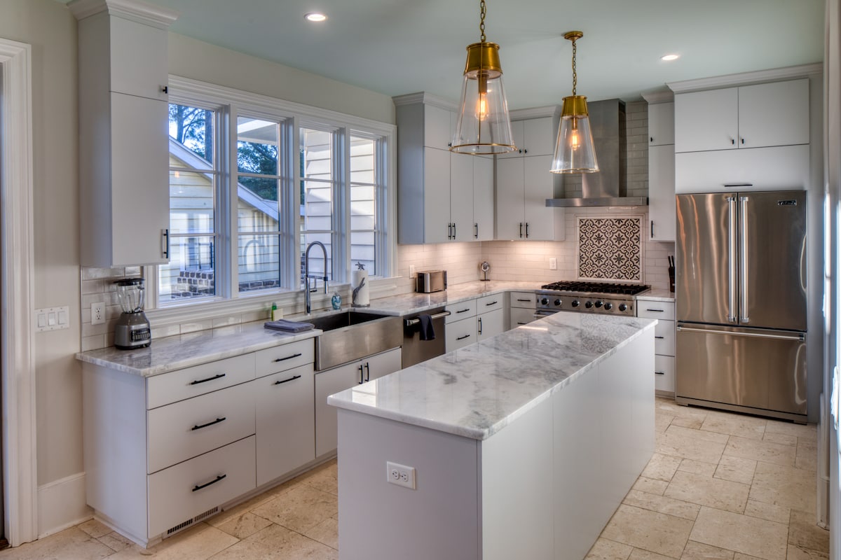 A modern kitchen features sleek cabinetry and a spacious central island with a marble countertop. Professional-grade stainless steel appliances are positioned along one wall, while decorative tile adds an elegant touch behind the stove. Large windows allow natural light to brighten the space.