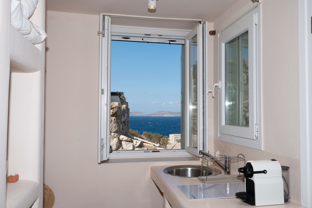 A kitchenette is visible, featuring a sink and a countertop. A window opens to reveal panoramic sea views, allowing natural light to fill the space. Neutral-toned walls complement the scenic landscape seen outside.