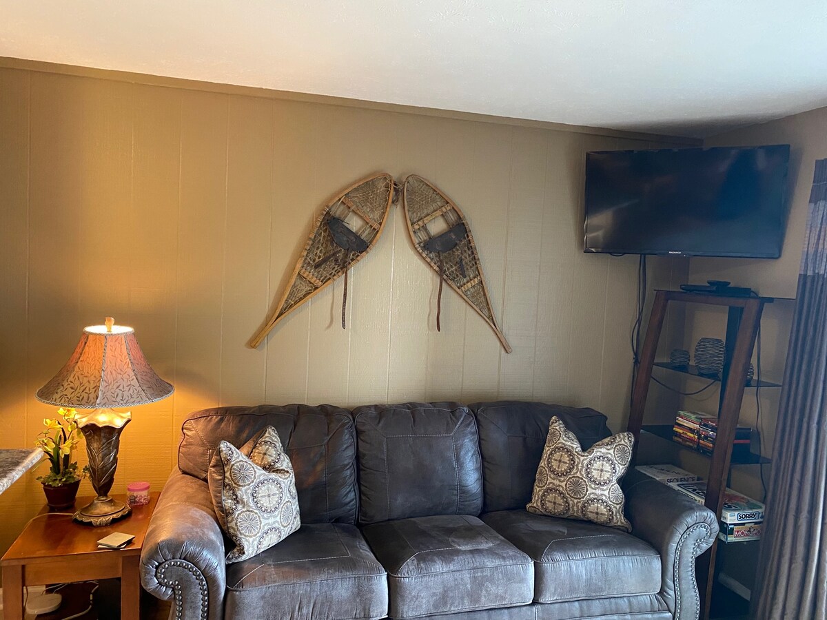 A comfortable seating area features a dark leather sofa adorned with patterned throw pillows. Above, a pair of vintage snowshoes is mounted on a light brown wall, while a flat-screen television is positioned nearby. A stylish lamp illuminates the space, providing a cozy ambiance.