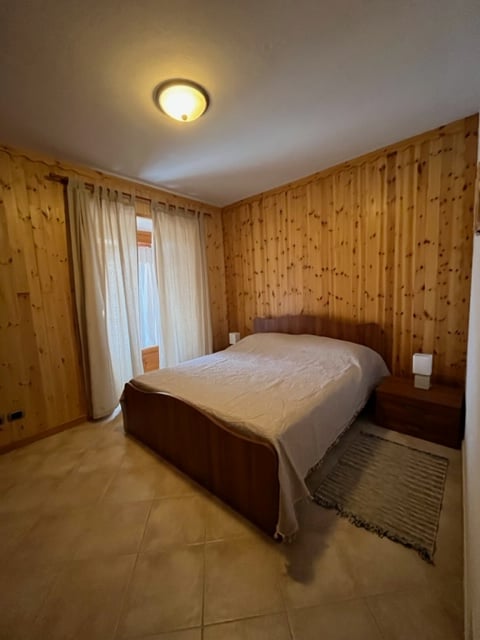 A spacious bedroom features wooden paneling on the walls, creating a warm ambiance. A large bed is positioned against the wall, complemented by matching nightstands on either side. Natural light filters through sheer curtains, enhancing the inviting atmosphere.