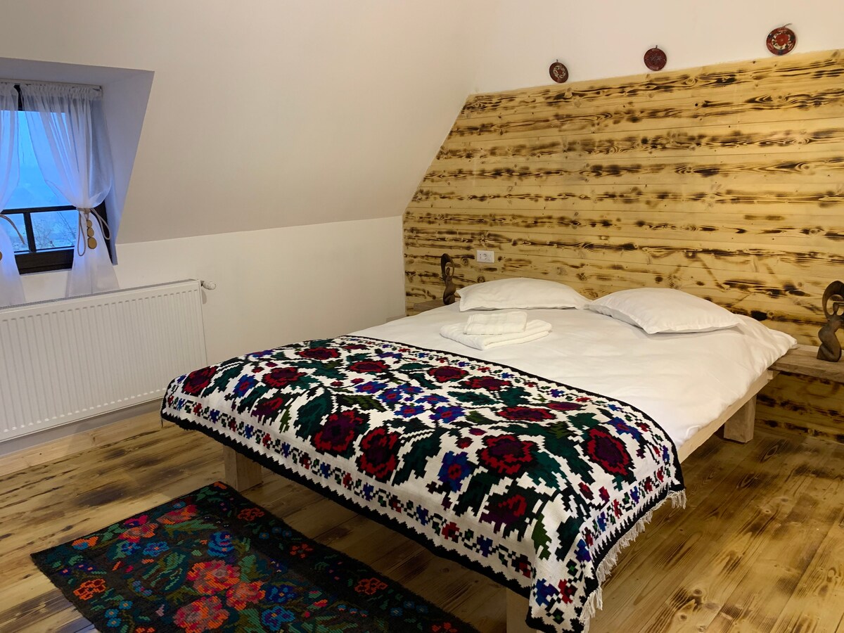 A warm bedroom features a wooden accent wall and a spacious bed topped with two white pillows. A colorful blanket with intricate floral patterns is draped over the bed. A patterned rug adds texture to the wooden floor, while a window allows natural light to enter.
