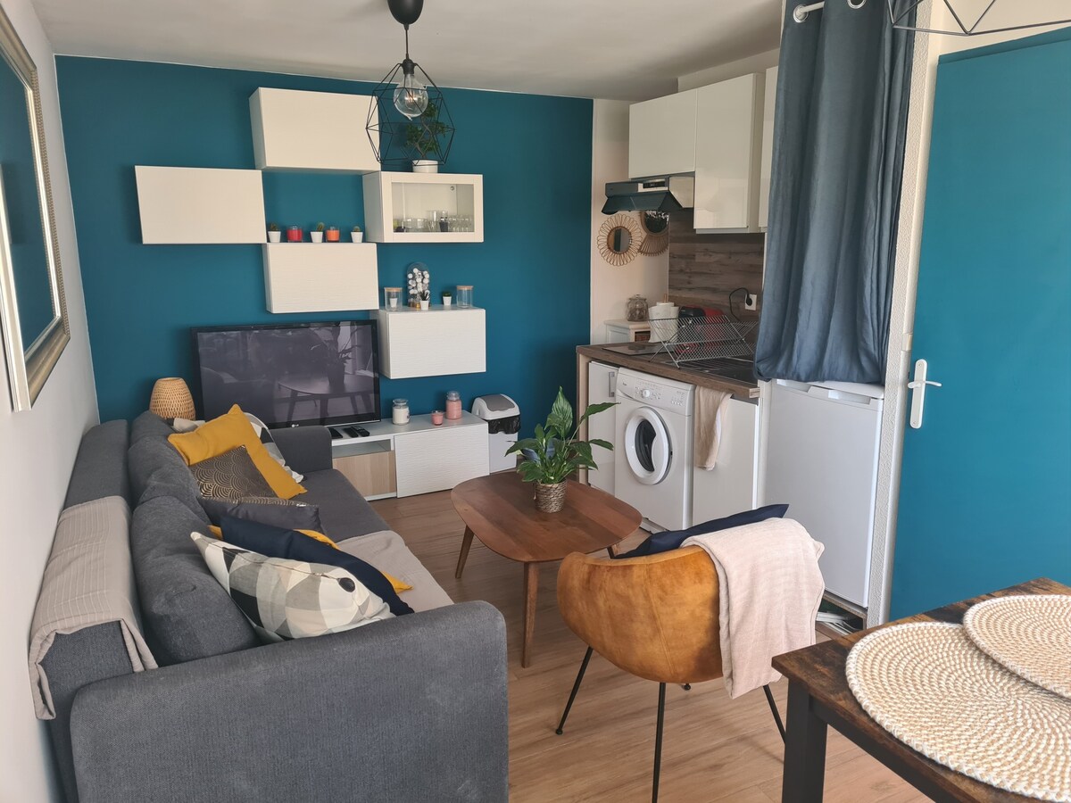 A cozy living area features a grey sofa adorned with colorful cushions. A small wooden coffee table sits in front, accompanied by a brown armchair. A TV is mounted on the wall, with shelves displaying decorative items. The space includes a kitchenette with a washing machine.