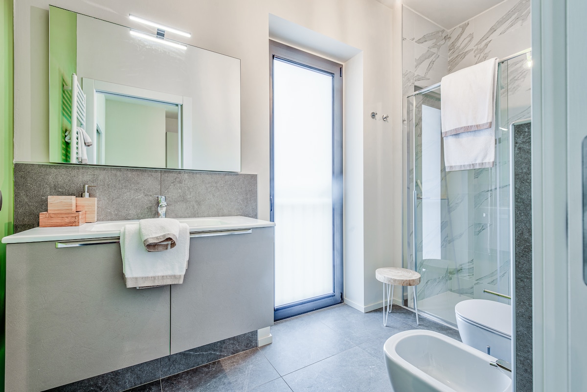 A modern bathroom features a spacious vanity with dual sinks, set against a sleek gray backdrop. A walk-in shower with glass doors sits beside a freestanding bathtub. Natural light streams through a large window, while neutral towels are neatly arranged on the counter.
