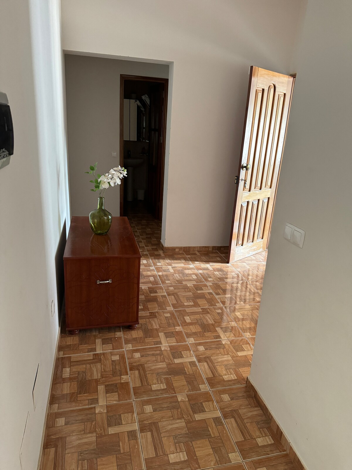 A well-lit hallway is observed, featuring a wooden cabinet with a subtle floral arrangement. The tiled floor reflects natural light, and a partially open door leads to another room, providing depth to the space.