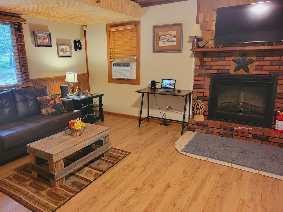 A cozy living area is presented with a dark, spacious sofa facing a brick fireplace. A rustic wooden coffee table is centrally positioned, and a small desk is set beside a window. Natural light enters through the blinds, enhancing the warm tones of the wood paneling.
