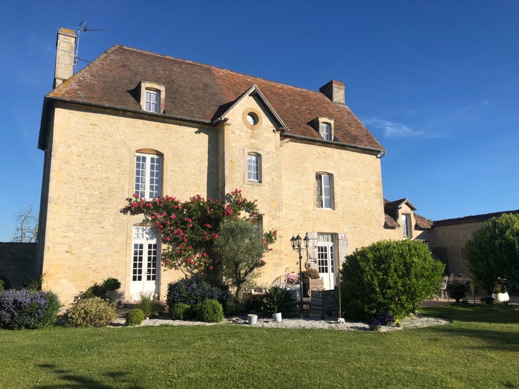 A charming two-story stone house is surrounded by lush greenery, featuring a well-maintained garden and flowerbeds. Large windows framed by climbing plants allow natural light to enter. A mix of shrubs and flowers adds color to the exterior, creating a serene outdoor space.