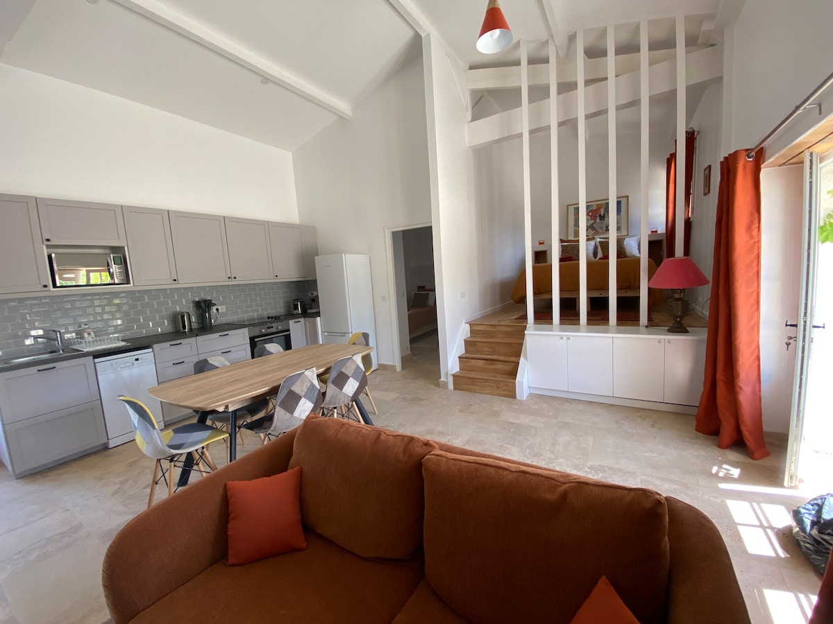 The open living space features a cozy sofa with decorative pillows, a dining table surrounded by stylish chairs, and a modern kitchen area furnished with sleek cabinetry. Soft light filters in through large glass doors, leading to a bedroom area highlighted by a softly elevated design.