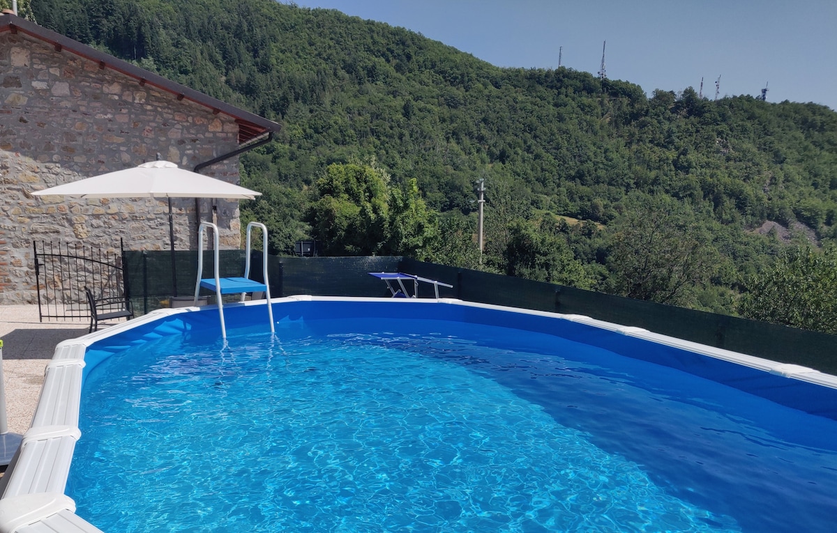 A circular above-ground swimming pool is seen, surrounded by a spacious terrace. Sun loungers and a large umbrella provide shaded seating. The backdrop features lush green hills, creating a serene outdoor environment.