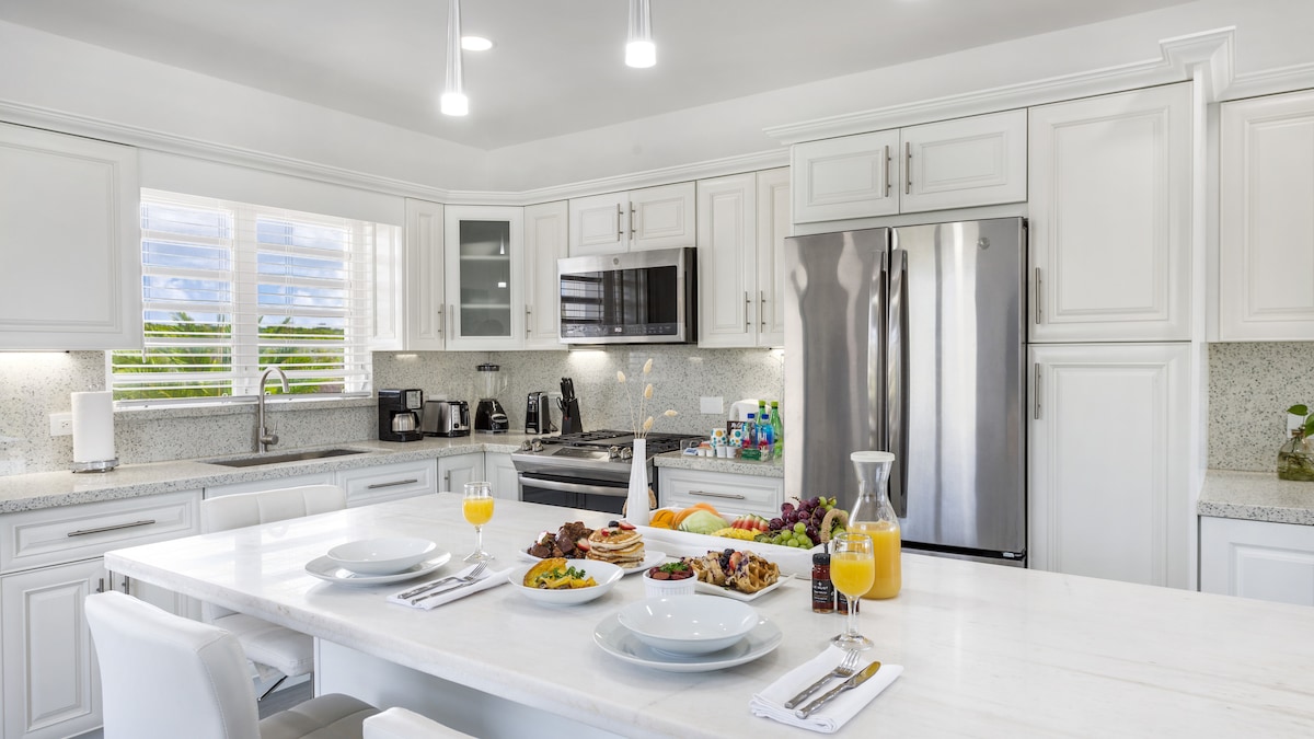 A modern kitchen is fitted with sleek white cabinetry and a large marble island. Bright lighting illuminates an array of breakfast items set on the island, including fresh fruit and beverages. Stainless steel appliances and a window offer a view of the outdoors.