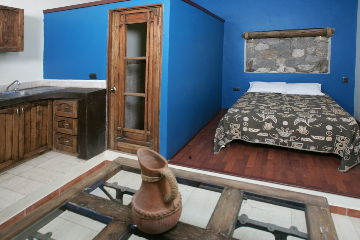 A loft-style space features a cozy sleeping area with a patterned bedspread against a blue wall. A wooden kitchen area displays rustic cabinetry. The layout allows for an open view of the room, with a wooden table at the forefront, complementing the overall design.