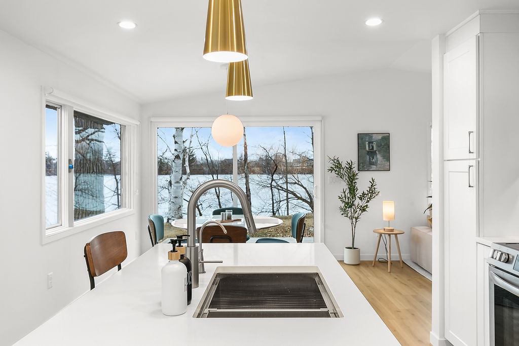 A modern kitchen features a large island with a sink, complemented by sleek, minimalist cabinetry. Bright light streams through expansive windows, highlighting a dining area with a round table and four chairs. The view shows greenery and a serene atmosphere outside.