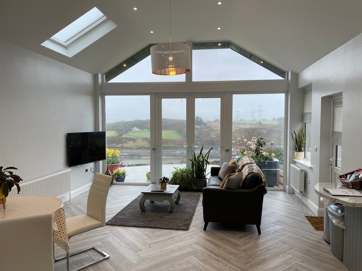 The interior of a bright living area features large glass doors that provide a view of the surrounding countryside. A comfortable sofa and coffee table are positioned on a patterned rug. A dining area with a circular table and chairs is also visible, along with decorative plants near the windows.