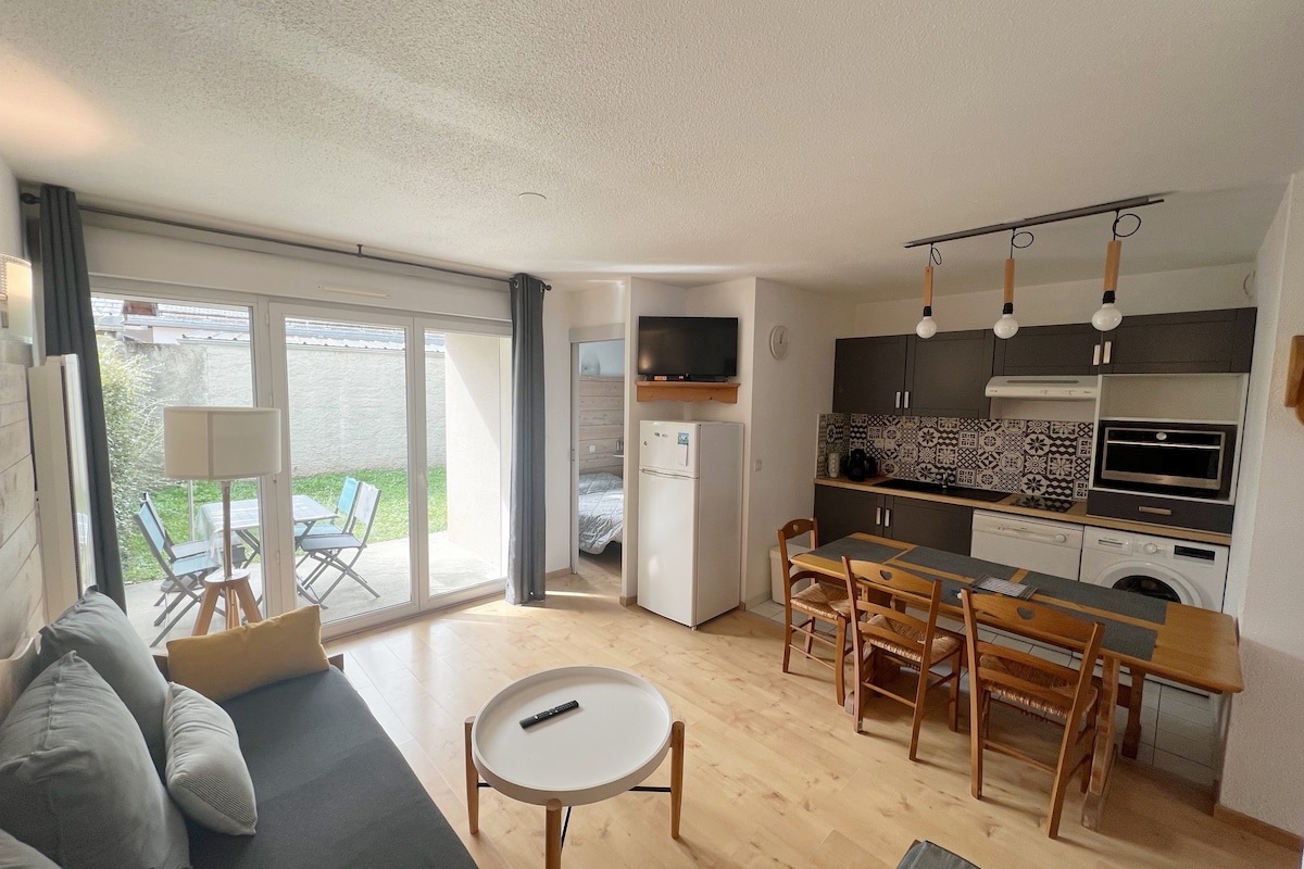 An open living area features a comfortable sofa, dining table, and a fully equipped kitchen with modern appliances. Large glass doors offer natural light and access to a green garden area. Decor includes light wood flooring and patterned backsplash tiles.