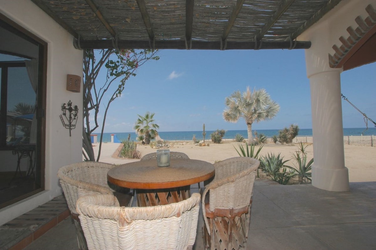 An outdoor seating area features a round table surrounded by four wicker chairs. The space is shaded by a thatched roof, offering views of the sandy beach and the Sea of Cortez in the distance.