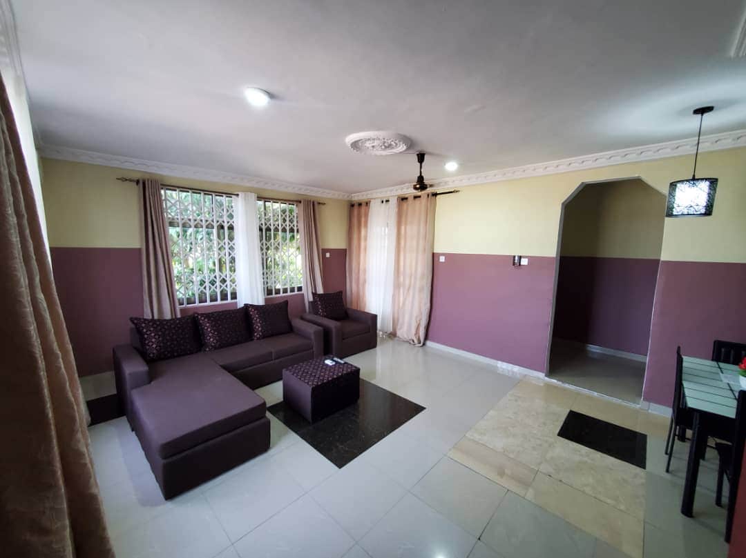 A spacious living area features a modern L-shaped sofa adorned with pillows. Natural light enters through a large window partially covered by light curtains. The floor is covered with glossy tiles, and a dining area is visible in the background, leading to a simple archway.