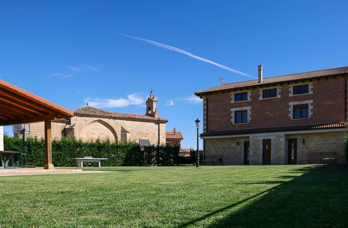 A spacious outdoor area presents well-maintained green grass, bordered by low hedges. The building features a two-story structure with a stone façade and multiple windows. A covered patio is visible on the left, along with a glimpse of a neighboring historical building.