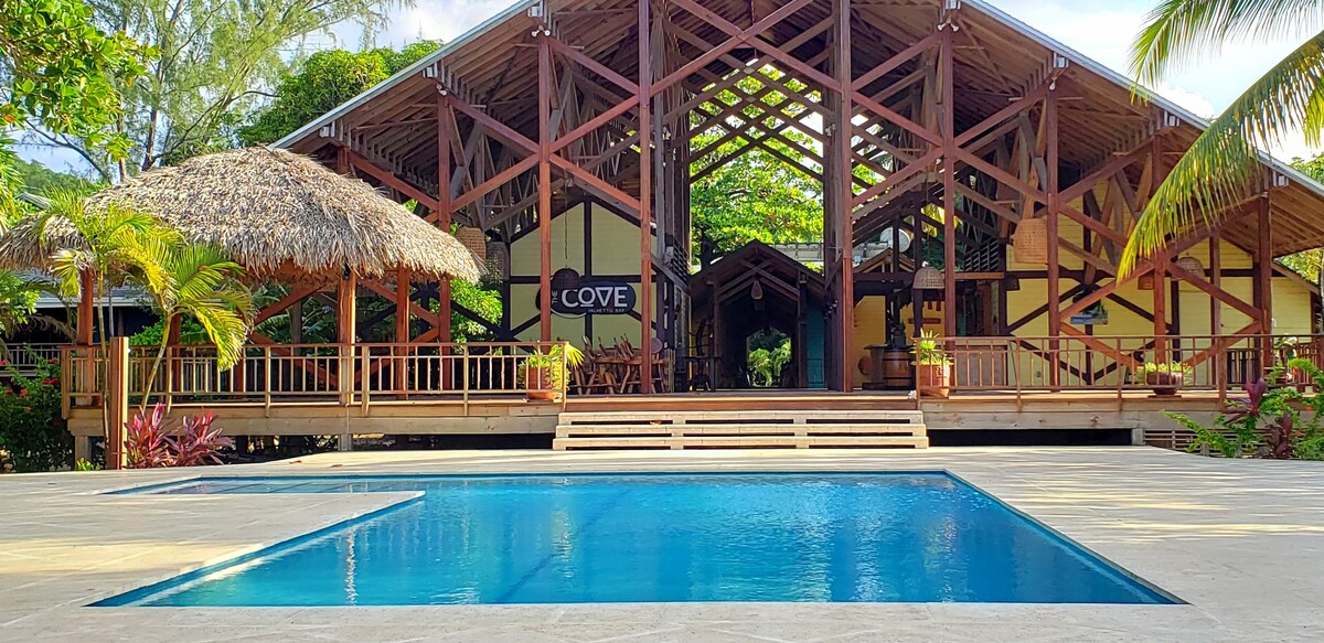 A clear view of an outdoor pool is presented, reflecting the surrounding lush greenery. The pool is bordered by a spacious wooden deck leading up to a large, open structure with thatched roofing and wooden beams, offering a relaxed gathering space.