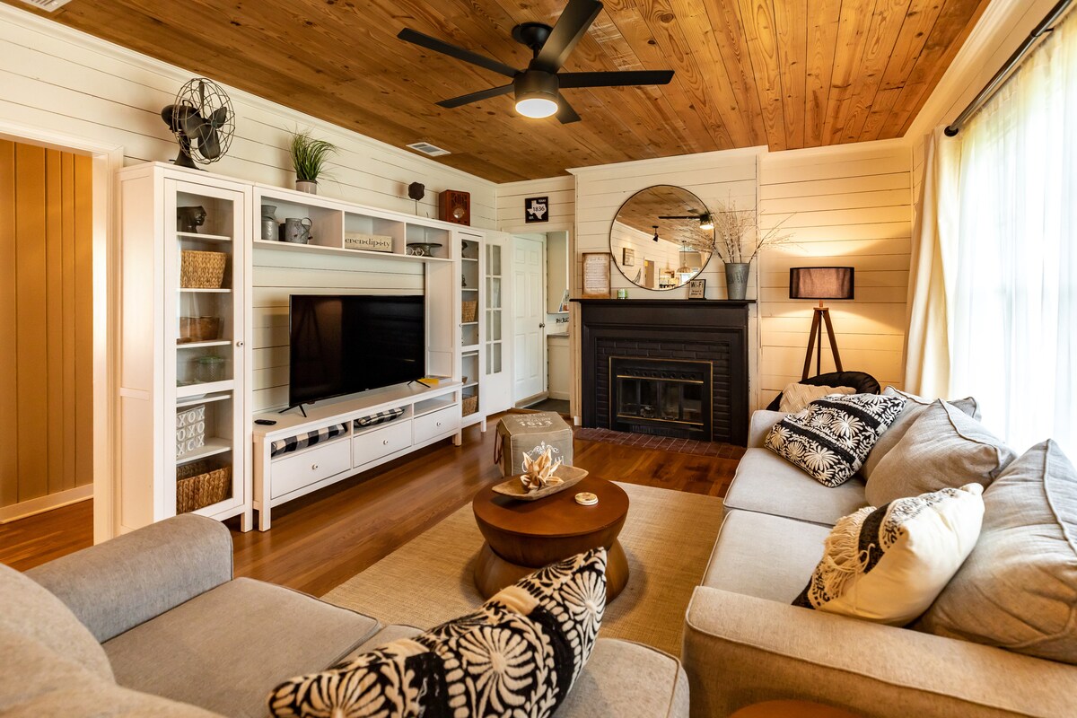 A cozy living room features a large sectional sofa, a round coffee table, and a white entertainment unit with a television. Wood paneling runs along the ceiling, complementing the warm tones of the hardwood floor. A fireplace serves as a focal point, framed by decorative accents.