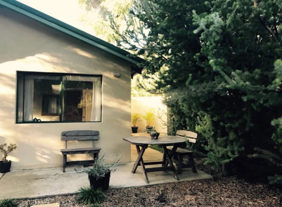 An outdoor area features a rustic wooden table surrounded by two matching benches. Potted plants add greenery to the setting, while trees provide shade and privacy. Natural light illuminates the space, which is adjacent to the cottage's entrance.