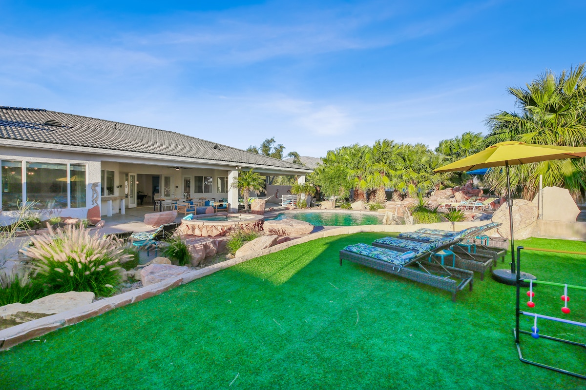 The outdoor area showcases a pristine saltwater pool surrounded by natural rocks and lush palm trees. Poolside loungers are positioned on green turf under a bright umbrella, with a built-in firepit and spa visible in the background next to the spacious house.
