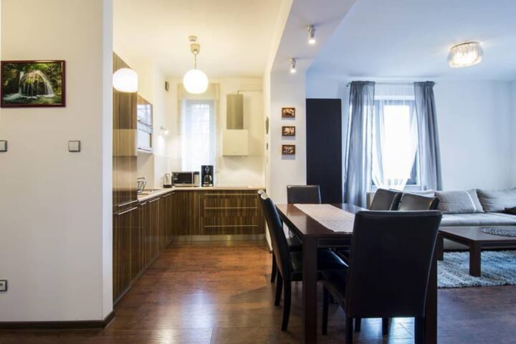 A modern kitchen area features sleek wooden cabinetry, complemented by stainless steel appliances. A dining table with dark chairs sits adjacent, while a comfortable seating area is visible, enhanced by natural light from a large window adorned with light gray curtains.