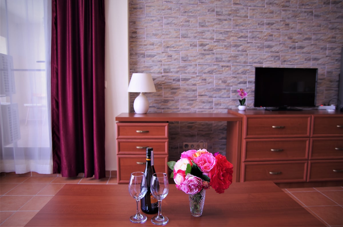 A wooden table is set with a bottle of champagne and two glasses, accompanied by a vibrant floral arrangement. Behind the table, a television is positioned on a wooden dresser, and a lamp provides additional lighting. Sheer curtains allow natural light to brighten the space.