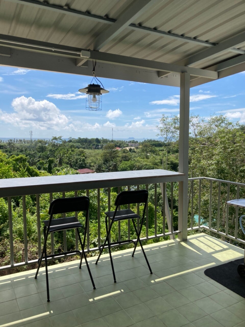 A covered terrace offers a scenic view of lush greenery and distant hills. Two black chairs are positioned at a high table, with sunlight illuminating the space. A pendant light hangs above, adding a touch of charm to this inviting outdoor area.